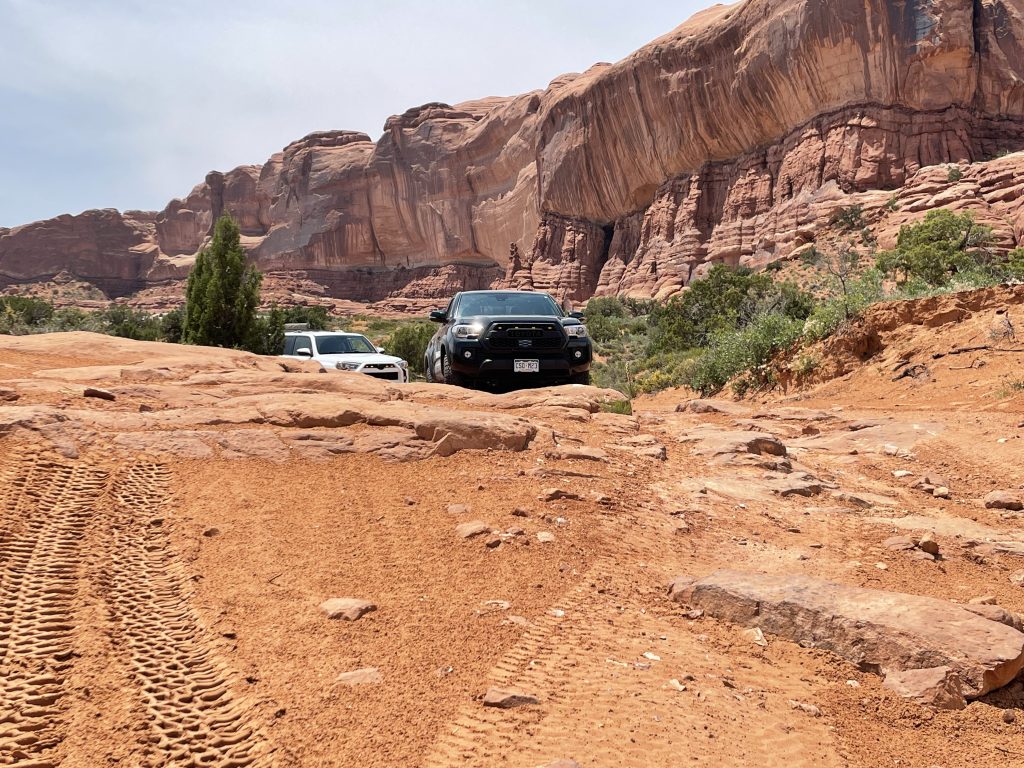 Black Tacoma truck about to descend slick rock ledges on Moab Wipeout Hill 4x4 trail followed by Toyota 4Runner.