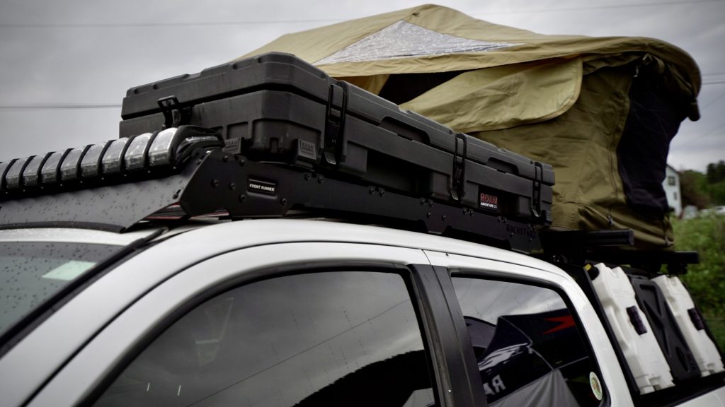 The top of a white Toyota Tacoma with overlanding accessories on the roof rack including a rooftop tent and Roam Adventure Co. 83L rugged case in black.