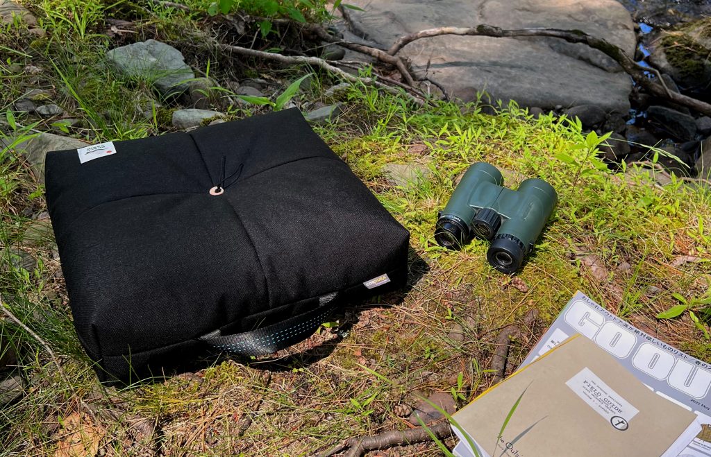 Peace Cabin Wanderer cushion outdoor floor pillow in black with other camping and overlanding accessories sitting on moss in the forest.