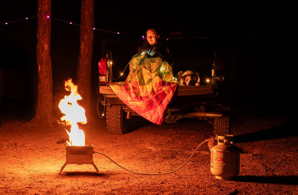 Woman and dog sitting on the back of a Toyota Tacoma with overlanding accessories like a colorful blanket and an ammo can fire pit.