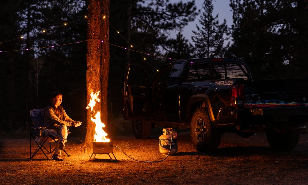 A woman sitting in a dark campsite, warming her hands next to a LavaBox Portable Campfire near a black Toyota Tacoma with string lights hung above her.