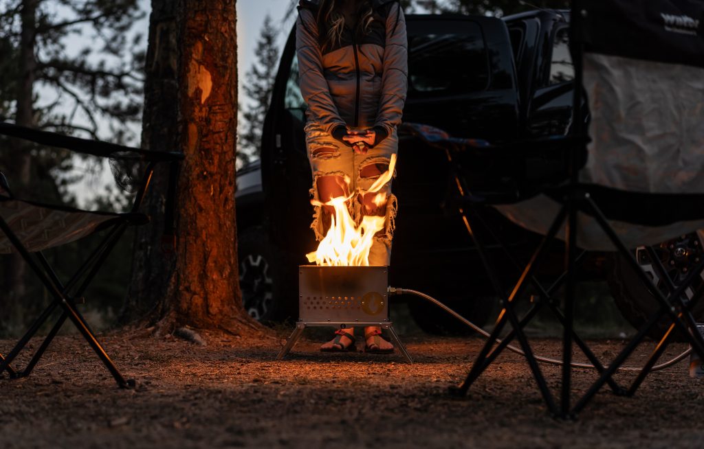 A woman warming her hands behind a LavaBox Portable Campfire Hekla stainless steel ammo can fire pit at a campsite.