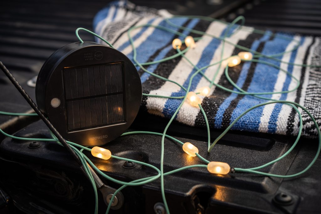 Luci Solar String lights next to the housing disk are sat on the tailgate of a Toyota Tacoma with a blue and white striped blanket.