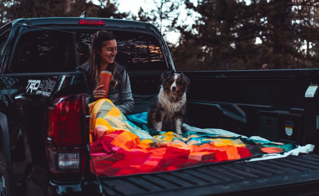 A woman in a grey sweater and an Australian shepherd sit in the bed of a black Toyota Tacoma at a campsite with a multicolored Rumpl outdoor blanket.