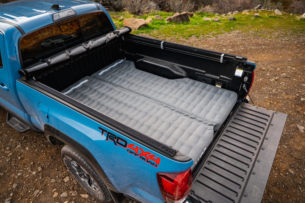 Bed of a blue Toyota Tacoma overlanding in the woods. The tailgate is down and a Luno Truck Bed Air Mattress is inflated in the bed.